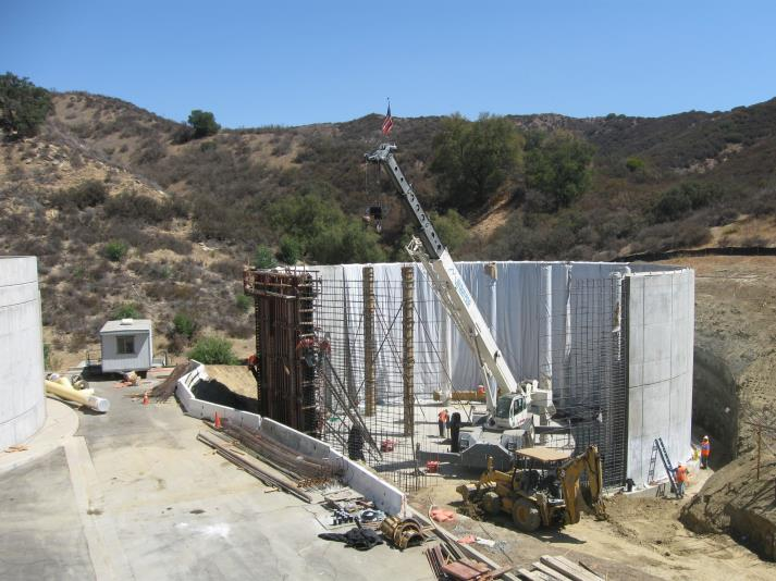 Construction site with a crane, workers, and machinery surrounded by hills and vegetation.
