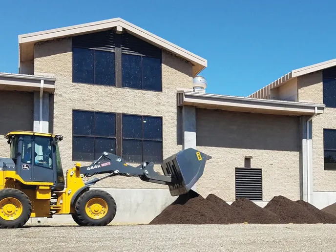 A yellow tractor is in front of a building, preparing to move piles of dark mulch on a gravel surface. Clear blue sky above.