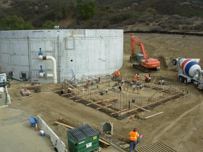 Construction site with workers, cement mixer, and an excavator near a large cylindrical concrete structure.