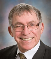 A smiling man with glasses, wearing a suit and tie, against a blurred background.
