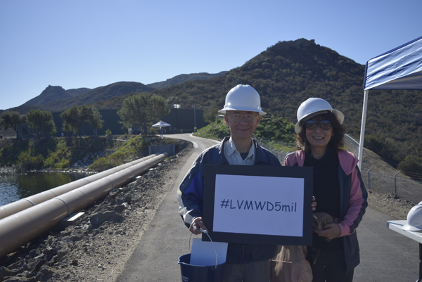 Two people in hard hats holding a sign with "#LVMWD5mil" outdoors near a mountain and pipeline.