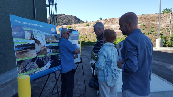 A group of people observe a presentation board about a five million gallon tank open house, outdoors in a sunny area.