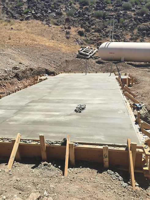 Concrete foundation slab with wood framing on a construction site, near a hill and a propane tank.