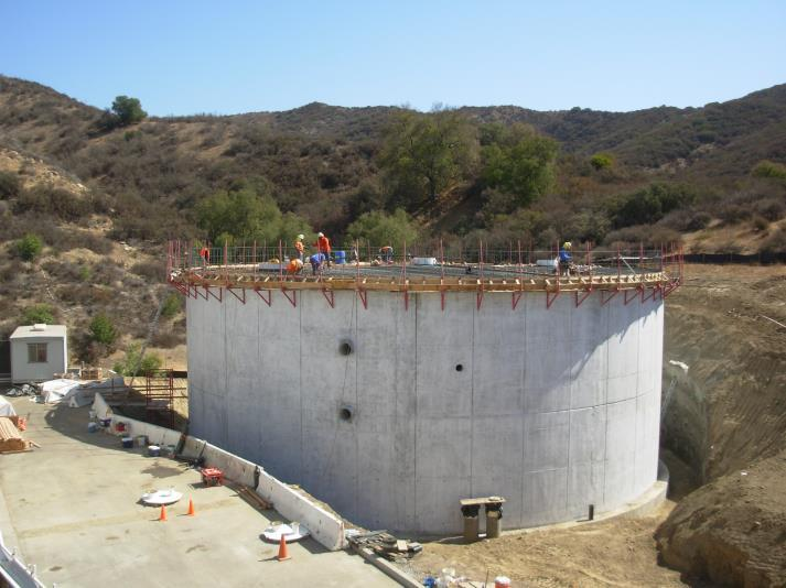 Large circular concrete structure under construction with workers on top, located in a hilly area.