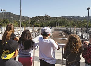 A group of people viewing a large outdoor facility with mountains in the background.