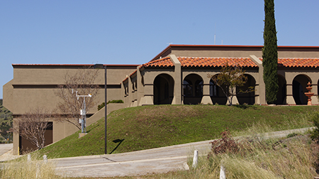 A building with a red-tiled roof, arches, a tree, and a lamp post on a grassy hill under a clear blue sky.