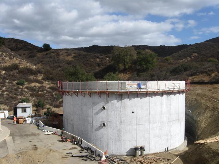 A large cylindrical concrete structure, possibly a water tank, in a hilly landscape with construction materials around.
