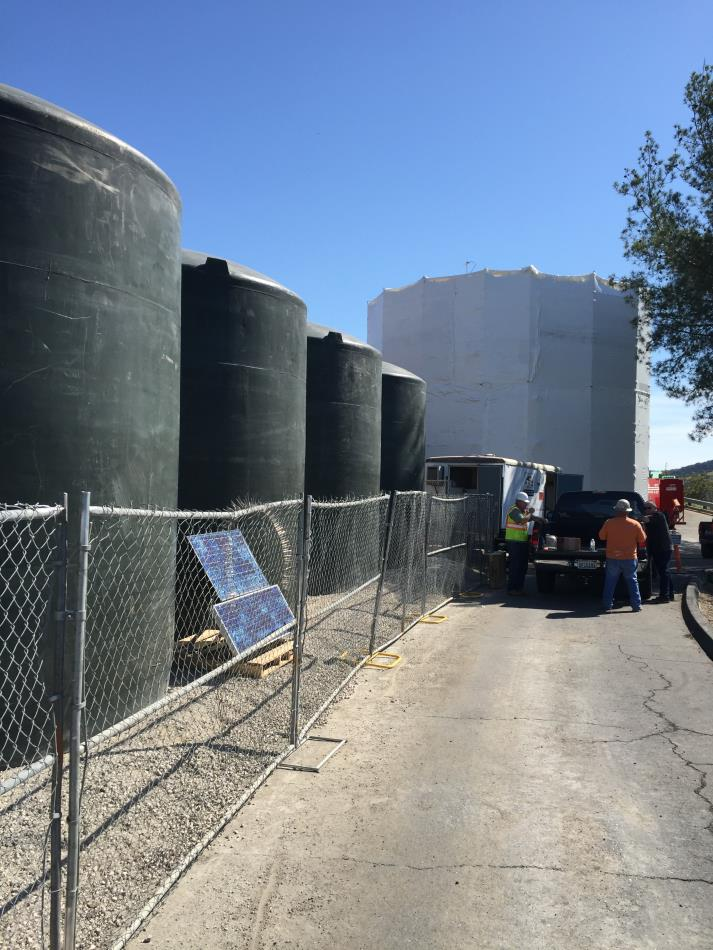 Large industrial tanks behind a chain-link fence, construction workers nearby, solar panels on the ground, clear blue sky.