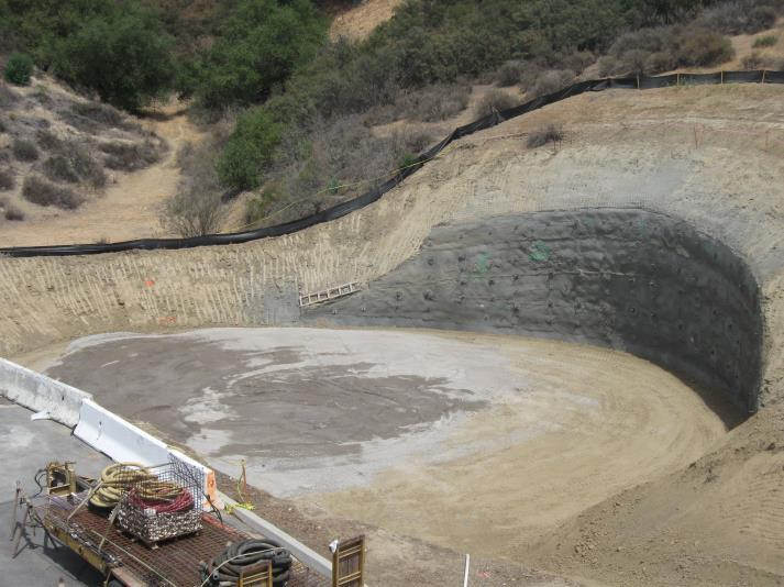 Construction site with a large excavated area surrounded by dirt and hills, and various construction materials in the foreground.