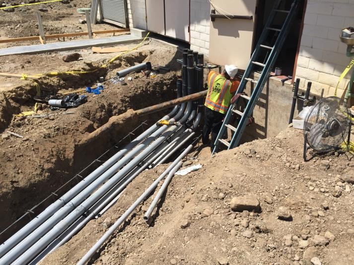 Construction site showing a worker in a safety vest and helmet near pipes, using a ladder beside a building entrance.