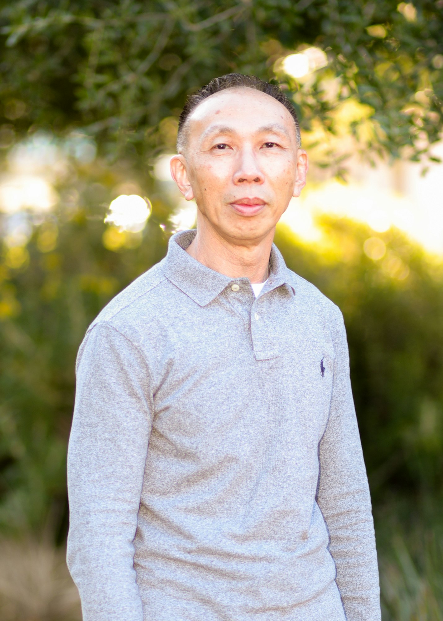A man in a gray polo shirt stands outdoors with greenery in the background, illuminated by soft, natural light.
