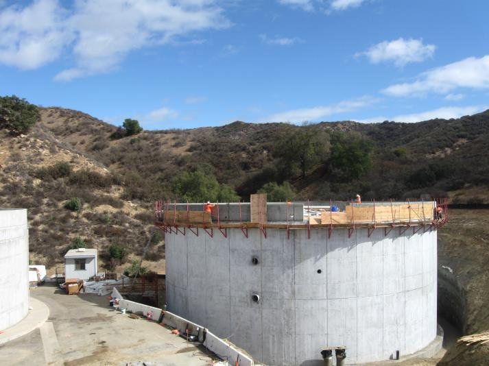 A large concrete structure under construction in a hilly landscape with a clear blue sky.