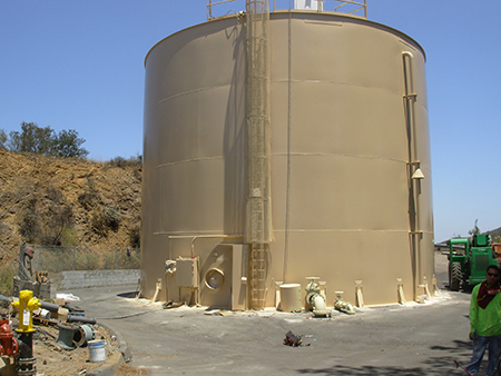 A large cylindrical water tank with a ladder, surrounded by piping and machinery, set in an outdoor industrial area.