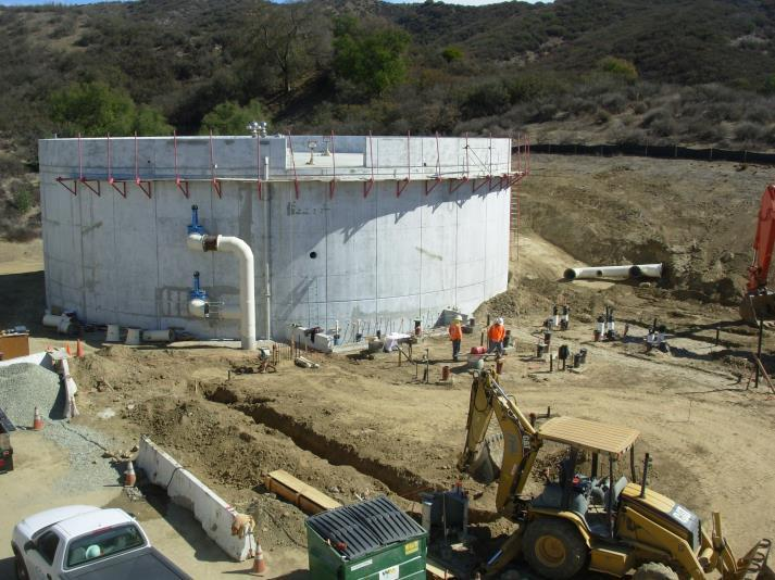 A construction site with a large circular concrete structure, workers, a backhoe, and surrounding hills.