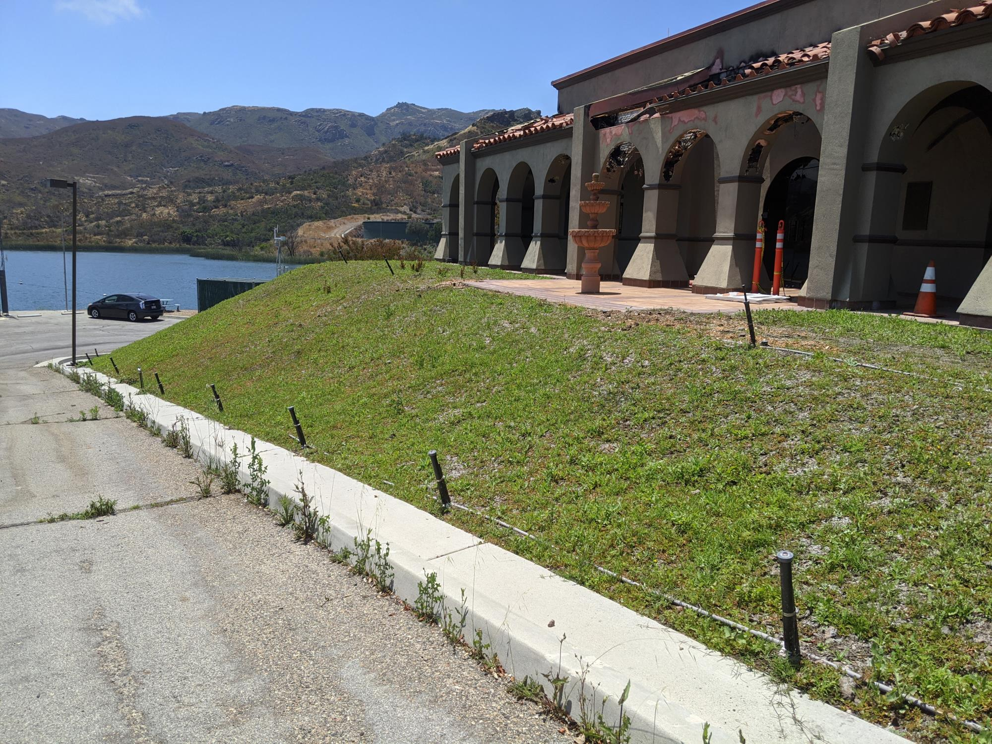 A lakeside building with arches, a fountain, grassy slope, car, and mountains in the background under a clear sky.