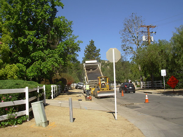 Roadwork scene with construction vehicles, cones, a car passing by, and traffic signs on a suburban street lined with trees.