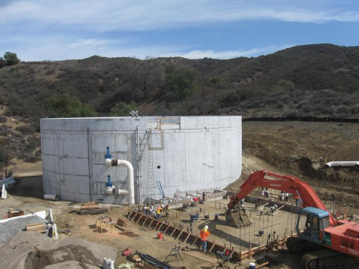 Construction site with a large concrete tank, excavator, workers, and building materials surrounded by hills.