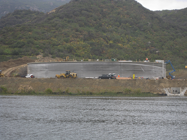 Large round structure under construction near a body of water, with vehicles and construction equipment around it.