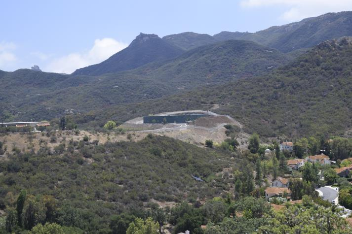 A hillside with houses and vegetation, set against a mountainous backdrop under a partly cloudy sky.