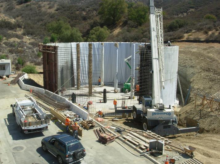 Construction site with workers, trucks, and a crane building a concrete structure next to a hillside.
