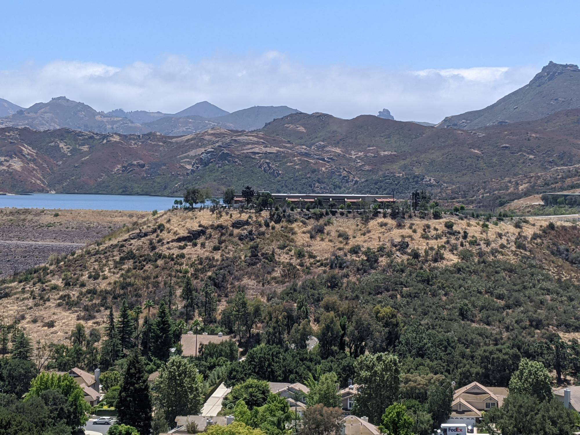 Houses and trees in the foreground, with hills, a lake, and mountains in the background under a blue sky.