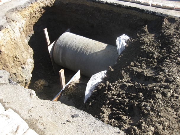 A large concrete pipe partially buried in a dug-out trench, supported by wooden stakes.