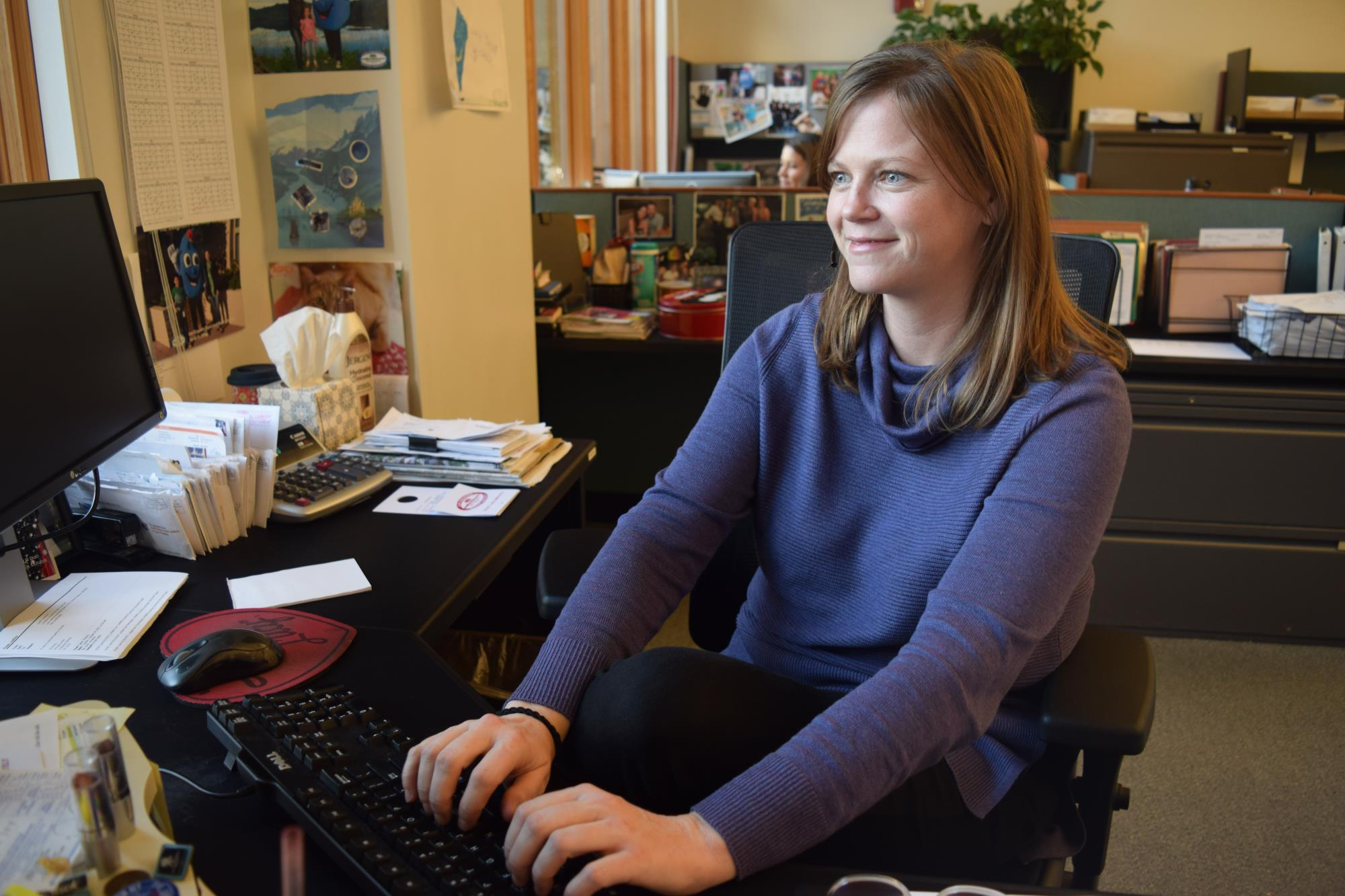 A woman in a blue sweater is smiling and typing on a keyboard in an office setting.