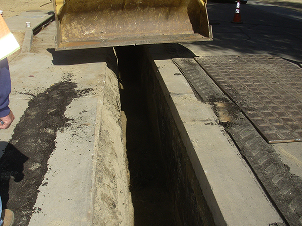 A trench on a road under a metal cover, with construction equipment and traffic cones nearby.