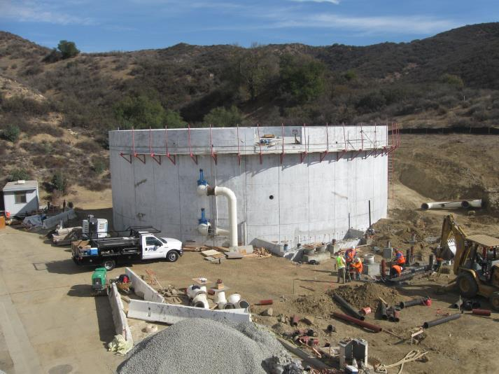 Construction site with a large circular structure, vehicles, pipes, and workers in a hilly area.