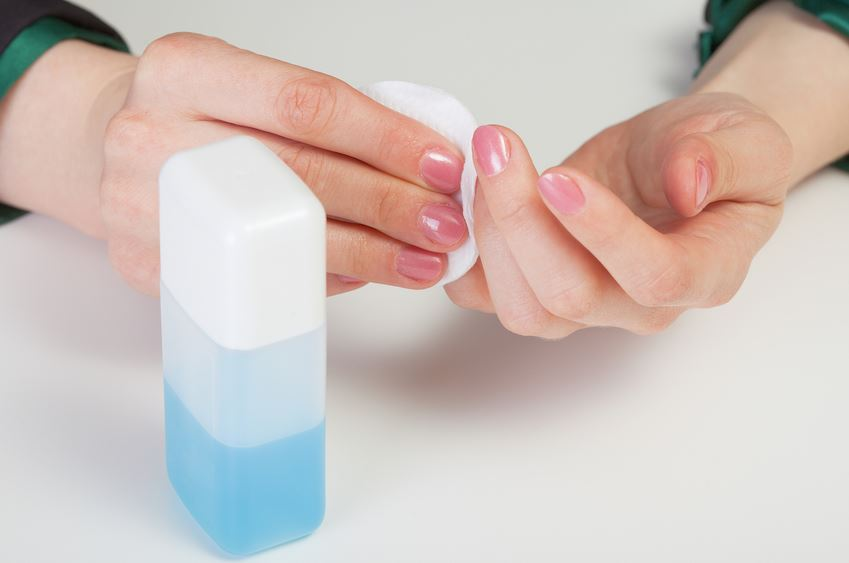 Hands using cotton pad and bottle of blue liquid for nail care or polish removal.