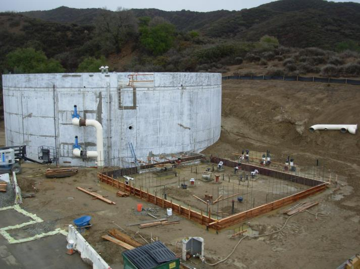A construction site with a large cylindrical structure, pipes, and a fenced-off area with equipment.