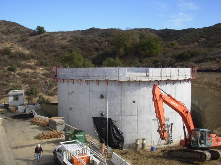 A construction site with a large cylindrical concrete structure, excavator, and surrounding equipment in a hilly area.