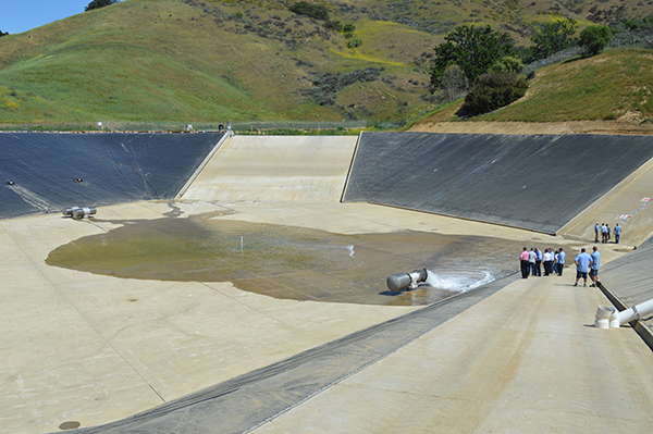 A group of people stands near a large concrete spillway with a small pool of water, surrounded by grassy hills.