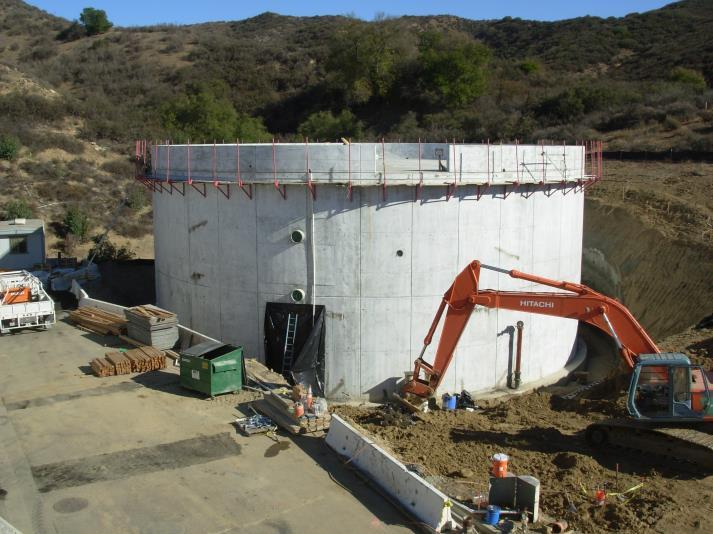 A construction site with a large concrete structure, an excavator, and materials, set against a hilly landscape.