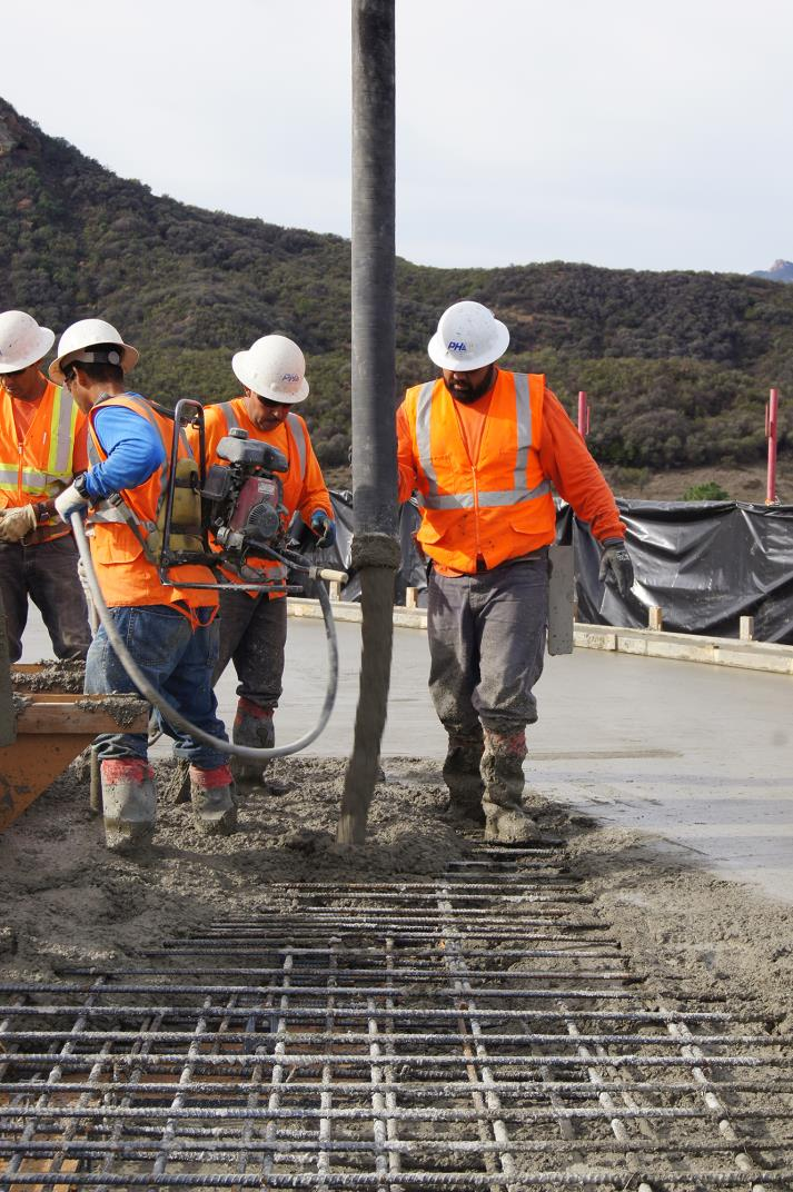 Construction workers pouring and leveling concrete on a rebar grid, wearing safety gear.