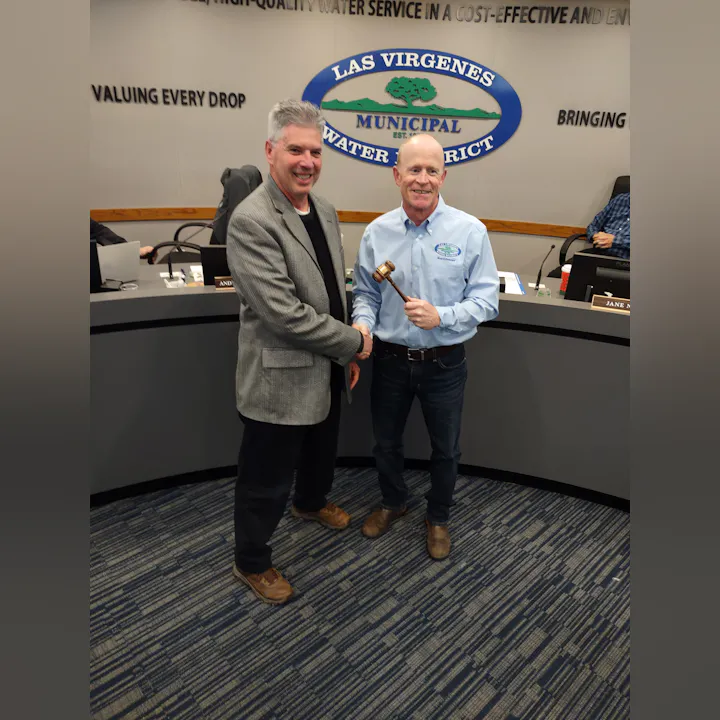 Two men shake hands in front of a water district sign; one holds a gavel, suggesting a leadership transition or meeting.