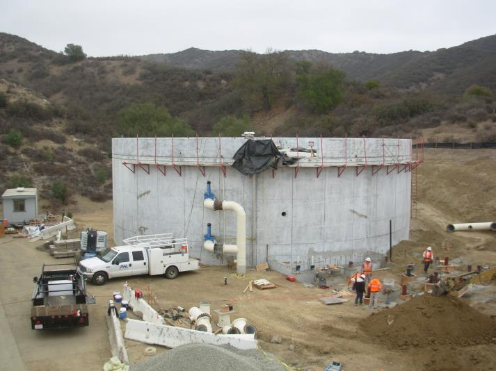 A construction site with a large concrete structure, vehicles, and several workers wearing safety vests in a hilly area.