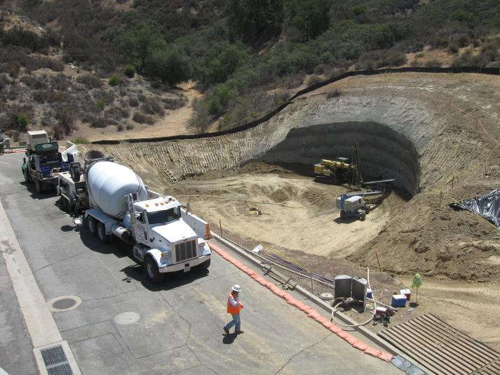 Construction site with a cement truck, excavated earth, construction workers, and machinery in a hilly area.