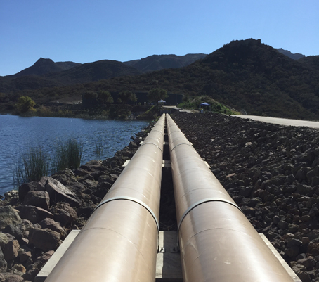 Pipes running parallel along a rocky path beside a body of water, with hills in the background under a clear blue sky.