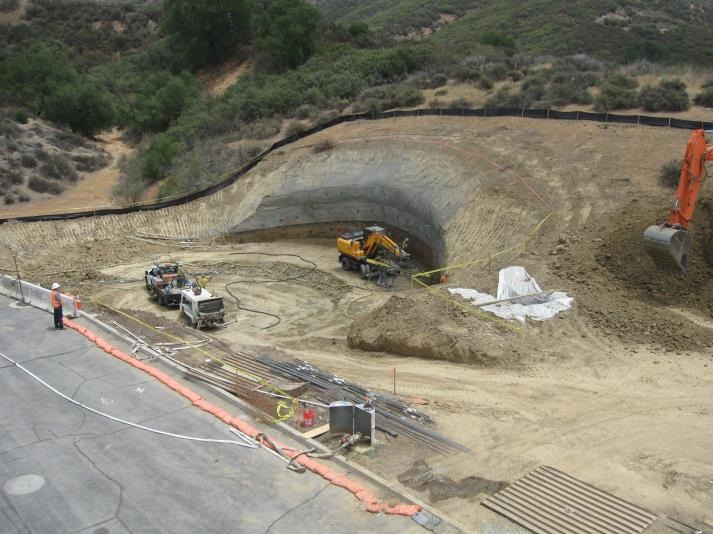 Construction site with excavation equipment, vehicles, and a cleared slope. Workers and materials are visible.