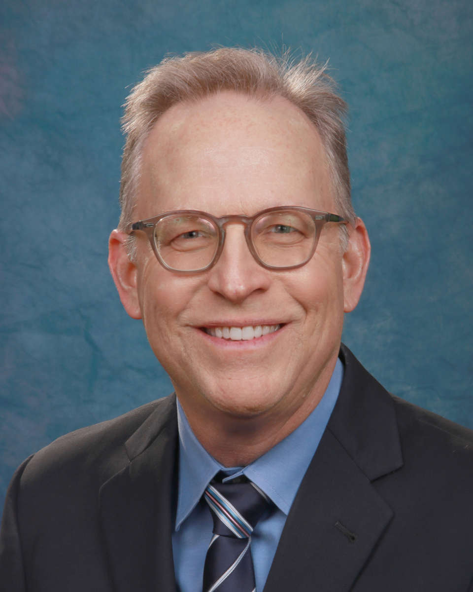 A man in a suit and glasses smiling with a blue background.