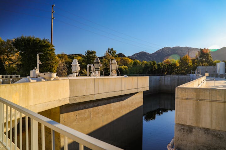 Concrete structure, possibly part of a water treatment facility or dam, with trees and mountains in the background under a clear sky.