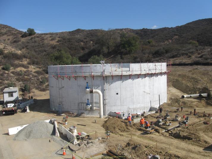 A large cylindrical concrete structure under construction on a hilly site, with workers and construction materials around it.