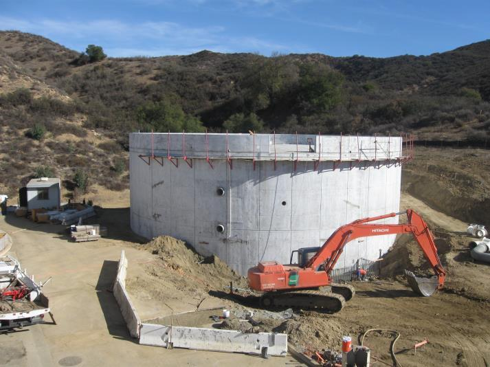 A construction site with a large circular concrete structure, an orange excavator, and surrounding hills.