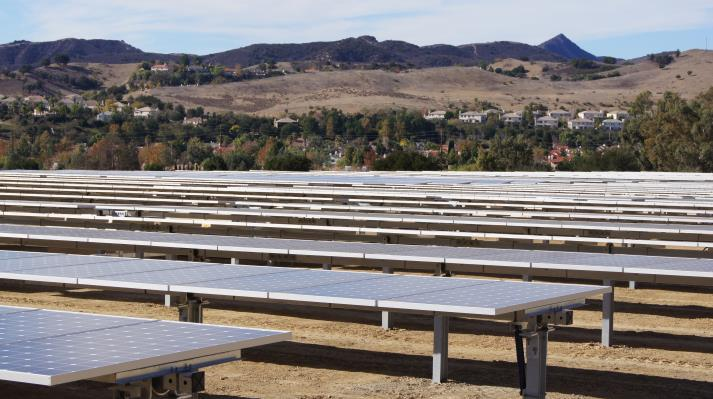 Rows of solar panels in a field with hills and houses in the background.