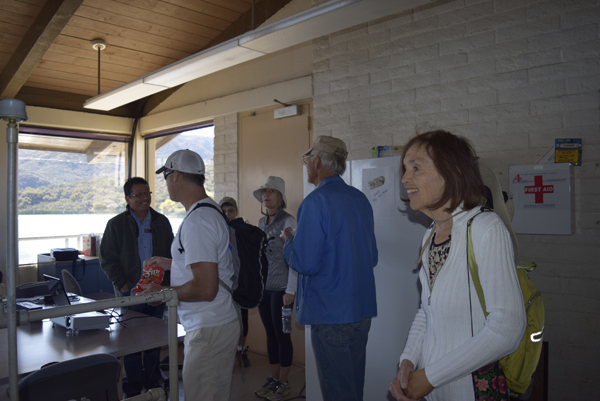 A group of people are gathered in a room with a view of mountains. There are tables, a fridge, and a first aid box visible.