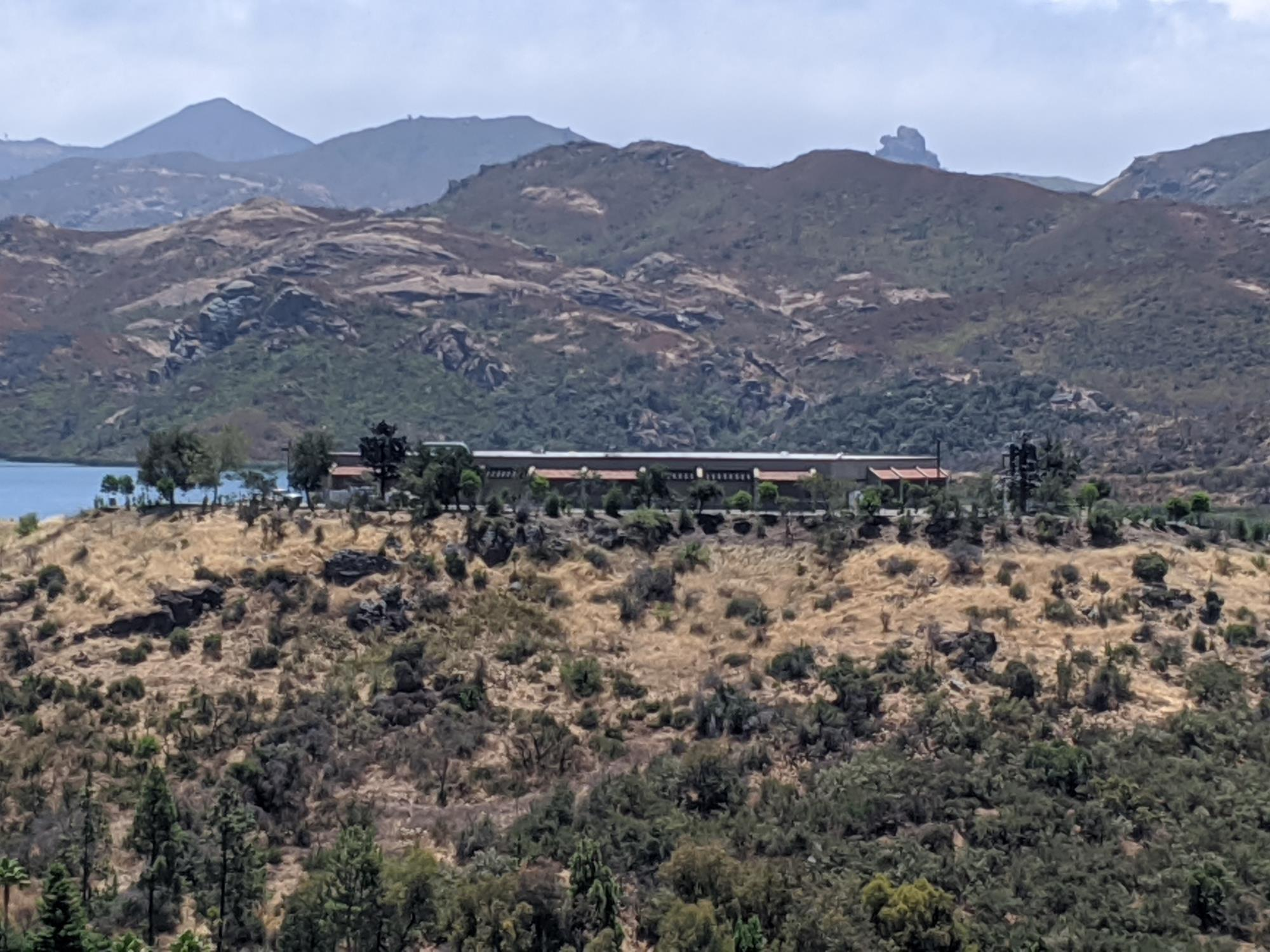 A building is nestled among trees on a hill, with mountains in the background and a body of water to the left.