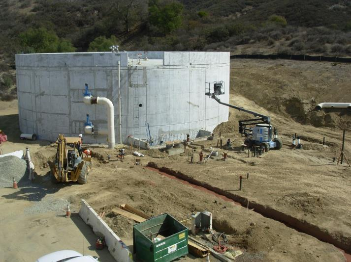Construction site with a large cylindrical concrete structure, earthmoving machinery, pipes, and scattered materials.