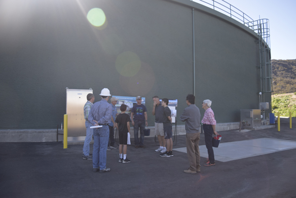 A group of people standing outdoors near a large industrial structure, discussing something.