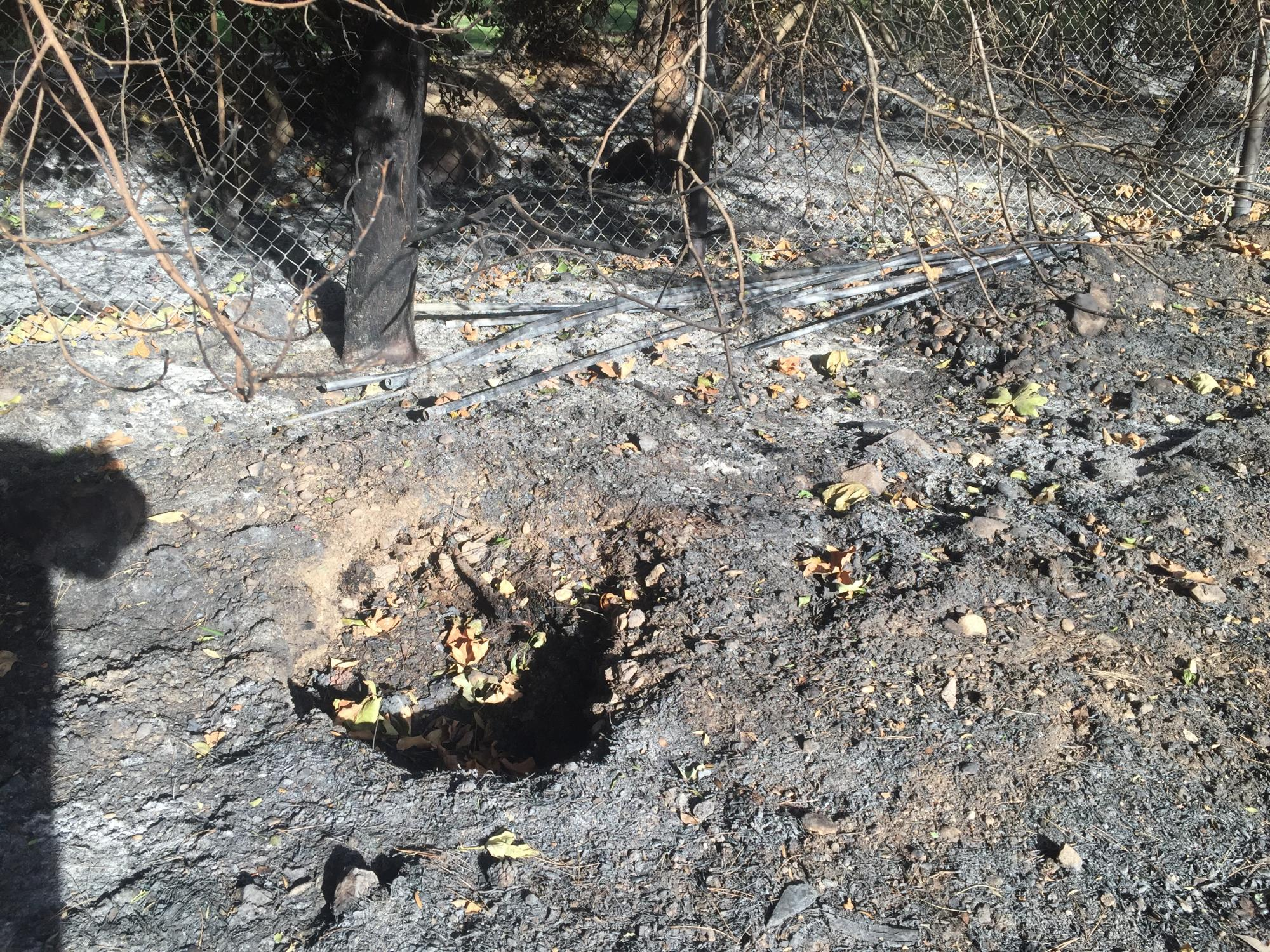 Scorched ground with some leaves, a hole, and a burnt fence, indicating fire damage.